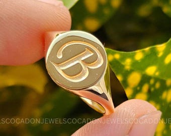 May include: A gold signet ring featuring a circular face engraved with the capital letter B. The ring has a polished metallic finish and a smooth, rounded band. The design is simple and consistent in color, showcasing the letter clearly on the face.