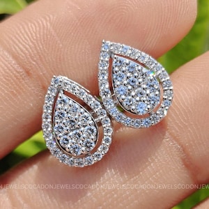 May include: A pair of silver-toned stud earrings, each featuring a pear-shaped cluster of small, clear, sparkling stones surrounded by a halo of additional stones. The text CADONJEWELSCO is visible in the lower right corner of the image.