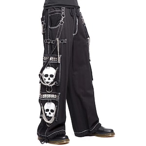 Op de afbeelding: Zwarte cargobroek met witte stiksels, kettingen en schedelpatches. De broek heeft meerdere zakken, metalen studs en een tailleband met trekkoord. Het ontwerp met wijde pijpen is zichtbaar en de broek wordt gedragen met zwarte laarzen.