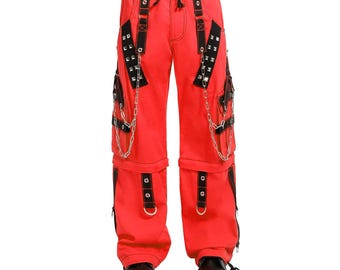 Op de afbeelding: Felrode broek met zwarte accenten, kettingen en zilveren studs. De broek heeft meerdere zakken, riemen en een taille met trekkoord. De onderkant van de broek is afneembaar. De broek wordt getoond met zwarte schoenen.