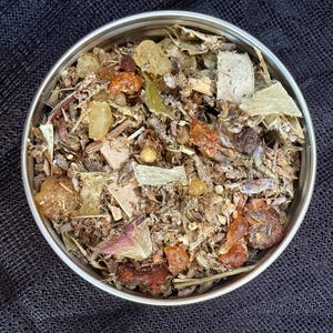 May include: A circular, silver-coloured tin filled with a mix of dried herbs, resins, and botanicals. The contents include pieces of amber, beige, and brown materials, with green and purple leaves. The tin sits on a dark, textured surface.