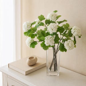 May include: A clear glass vase holds a bouquet of white snowball flowers with green leaves. The vase sits on a white surface next to a closed book and a small, beige stone sculpture. The background is a neutral color.