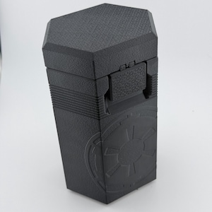 May include: A black hexagonal storage container with a textured surface and a hinged lid. The front panel features a debossed circular emblem with a geometric starburst pattern. The container has a secure latch mechanism and a tapered body design.