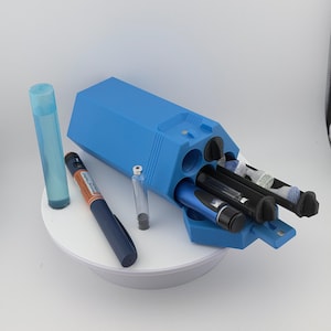 May include: A blue medical device organiser with multiple compartments, holding several insulin pens and vials. A blue pen and a clear vial are placed next to the organiser. A clear, cylindrical container is also present.