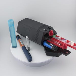 May include: A black medical organiser with red trays, containing medical supplies such as syringes and vials. A blue pen-like device, a clear vial, and a blue cylindrical container are also present. The items are arranged on a white surface.