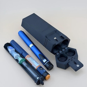 May include: A black, 3D-printed, hexagonal-shaped carrying case with a hinged lid, containing a blue Novolog FlexPen insulin injector and a separate black case holding a clear syringe with a green-capped needle and a blue-capped needle.