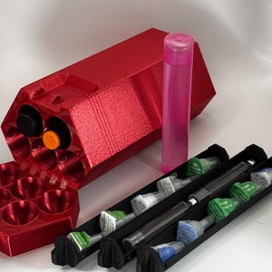 May include: A red, hexagonal container with two black and orange circular openings. A pink, cylindrical tube stands upright. Several syringes are in black trays, and a red tray with circular openings is also present.