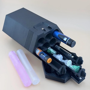 May include: A black plastic organizer with multiple cylindrical slots containing two insulin pens, three needle tips, and one glass vial. Two separate translucent cylindrical tubes, one light pink and one clear, are positioned next to the organizer.