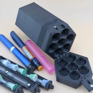 May include: A black hexagonal storage case with seven circular compartments, accompanied by a blue insulin pen, a NovoLog FlexPen, a pink protective pen case, and a black tray containing several medical needle tips with green and blue caps.