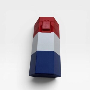 May include: A hexagonal container featuring a vertical color block design with a red top section, a white middle section, and a blue bottom section. The top red portion includes a small, square-shaped latch mechanism centered on the front face.