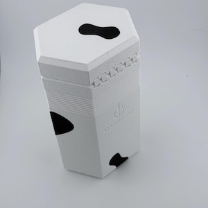 May include: A white hexagonal container with a textured surface and black organic-shaped spots. The lid features a hinge mechanism on one side and a black spot on top. A small embossed logo is visible on the side with the text DECKOFTHEDAY.