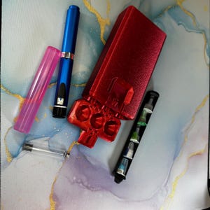 以下が含まれることがあります： 赤色の長方形ケース、青いペン、ピンクのチューブ、小さなカラフルなバイアルがいくつか入った黒い容器など、さまざまなアイテムのコレクション。アイテムは、青と金の模様の表面に配置されています。