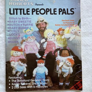 May include: A softcover book titled Xavier Roberts Presents Little People Pals, Collector's Edition #7546 Plaid. The cover features a man in a cowboy hat surrounded by several soft-sculpture dolls with yarn hair and various outfits, plus text listing doll names and book features.