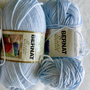 May include: Several skeins of light blue Bernat Satin yarn are arranged on a white surface. The yarn is labeled as 100% acrylic, worsted weight, and 3.5 oz/100g. The label also mentions a free pattern.