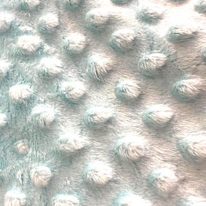 Puede incluir: Primer plano de una manta azul claro con una superficie texturizada. La manta tiene una textura suave y con bultos con mechones redondeados en relieve, creando una apariencia acogedora. La tela parece suave y c&aacute;lida.