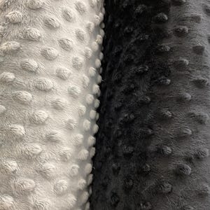 Puede incluir: Primer plano de dos rollos de tela de felpa. Un rollo es gris claro y el otro gris oscuro. Ambas telas tienen una superficie texturizada y con bultos, creando una apariencia suave y t&aacute;ctil. La tela probablemente se utiliza para mantas u otros art&iacute;culos acogedores.