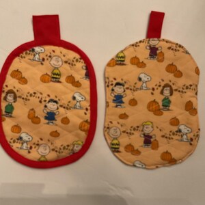 May include: Two quilted pot holders with a red trim and a red loop. The pot holders have a light orange background with a pattern of pumpkins and cartoon characters. One is oval, the other is a rounded shape.