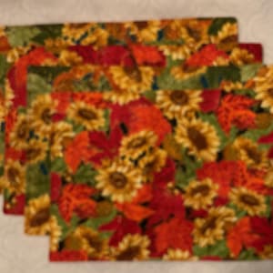 May include: Four rectangular placemats with a vibrant floral pattern. The design features sunflowers, red and orange autumn leaves, and green foliage. The placemats are arranged in a stack, showcasing the repeating pattern.
