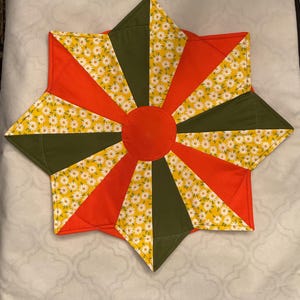 May include: A star-shaped quilted trivet with a central orange circle and radiating segments of orange, olive green, and yellow daisy-patterned fabric. The trivet is designed with a geometric pattern and a stitched edge.