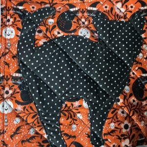 Puede incluir: Un agarrador de cocina acolchado cuadrado con un fondo de tela naranja que presenta un patrón repetitivo de gatos negros, calabazas blancas y calaveras. Una gran aplicación de gato negro con lunares blancos está cosida en el centro.