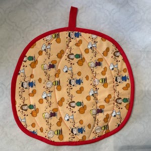 May include: A pumpkin-shaped pot holder with a red trim and loop. The fabric features a repeating pattern of cartoon characters, pumpkins, and autumn leaves on a light orange background. The pot holder is designed for kitchen use.