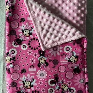 Puede incluir: Manta rosa para beb&eacute; con un dise&ntilde;o de Minnie Mouse, flores y c&iacute;rculos. La manta tiene un reverso rosa suave y texturizado. El dise&ntilde;o incluye a Minnie Mouse en varias poses, con un tema floral y de lunares.