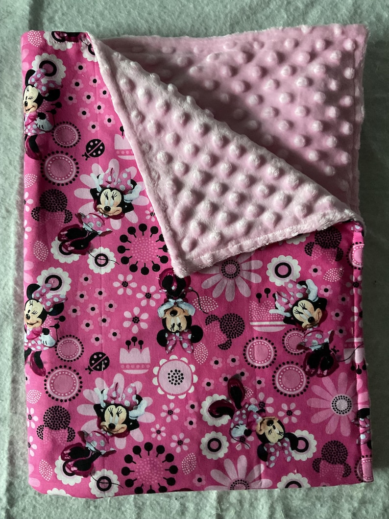 Puede incluir: Manta rosa para beb&eacute; con un dise&ntilde;o de Minnie Mouse, flores y c&iacute;rculos. La manta tiene un reverso rosa suave y texturizado. El dise&ntilde;o incluye a Minnie Mouse en varias poses, con un tema floral y de lunares.