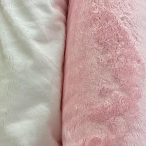 Puede incluir: Primer plano de dos rollos de tela de felpa. Un rollo es blanco suave y esponjoso, y el otro es rosa claro. La tela tiene un aspecto texturizado y aterciopelado, lo que sugiere que es suave al tacto. Ideal para mantas u otros proyectos textiles.