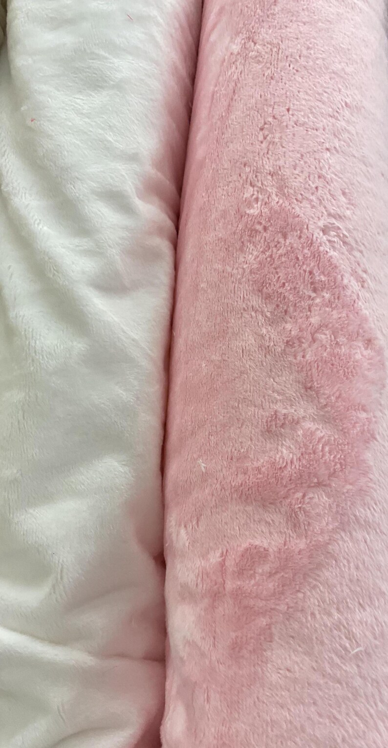 Puede incluir: Primer plano de dos rollos de tela de felpa. Un rollo es blanco suave y esponjoso, y el otro es rosa claro. La tela tiene un aspecto texturizado y aterciopelado, lo que sugiere que es suave al tacto. Ideal para mantas u otros proyectos textiles.