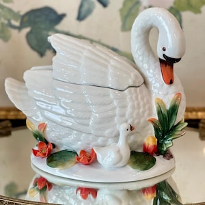 May include: A white ceramic swan-shaped lidded tureen featuring a sculpted swan with an orange beak and a smaller cygnet at its base. The base is decorated with green lily pads and small orange flowers, creating a detailed, three-dimensional scene.