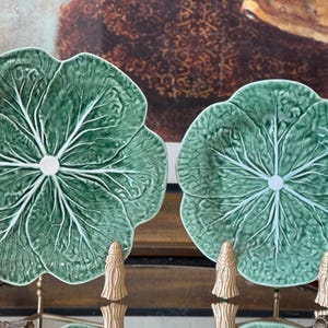 May include: Two green ceramic plates shaped like cabbage leaves with prominent white veining and scalloped edges. Each plate features a glossy finish and a central white circular detail, displayed upright on small gold-toned metal stands.