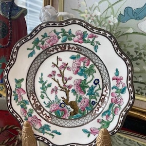 May include: A white porcelain plate with a scalloped edge and a brown border. The plate features a floral design with pink and teal flowers, green leaves, and a brown tree. The plate is supported by a gold-colored stand.