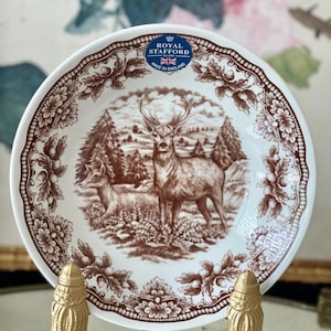 May include: A white ceramic plate featuring a brown transferware print of a stag and a doe in a forest setting. The rim is decorated with a brown pattern of oak leaves and acorns. A blue and white Royal Stafford label is visible at the top.