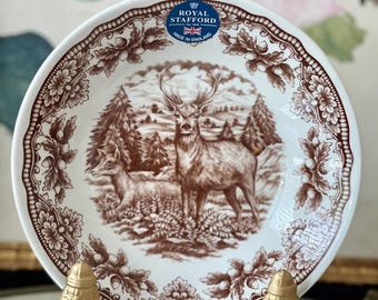May include: A white ceramic bowl featuring a brown transferware print of a stag and a doe in a forest setting. The rim is decorated with a repeating pattern of oak leaves and acorns. A blue and white Royal Stafford Made in England label is present.