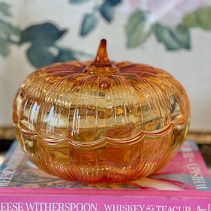 May include: A translucent amber-colored glass pumpkin-shaped lidded dish with vertical ribbed detailing and a small pointed stem at the top. The dish is resting on two stacked pink books with the text Whiskey in a Teacup printed on the spines.