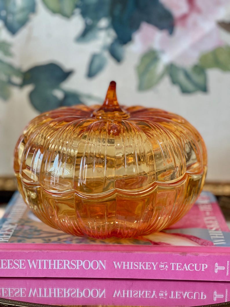 May include: A translucent amber-colored glass pumpkin-shaped lidded dish with vertical ribbed detailing and a small pointed stem at the top. The dish is resting on two stacked pink books with the text Whiskey in a Teacup printed on the spines.