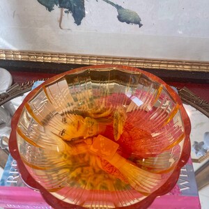 May include: A translucent amber-colored glass bowl with a scalloped rim and vertical ribbed detailing along the interior walls. The bowl features a wide, circular opening and a pedestal base, resting atop a pink book with visible white text.