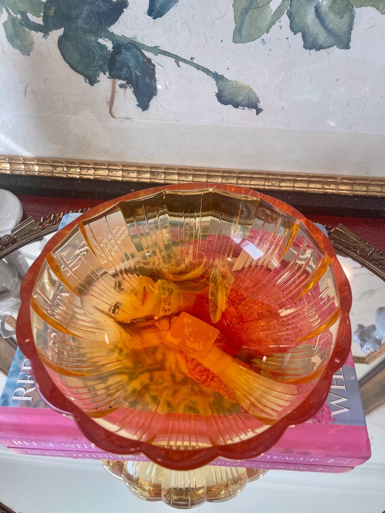 May include: A translucent amber-colored glass bowl with a scalloped rim and vertical ribbed detailing along the interior walls. The bowl features a wide, circular opening and a pedestal base, resting atop a pink book with visible white text.