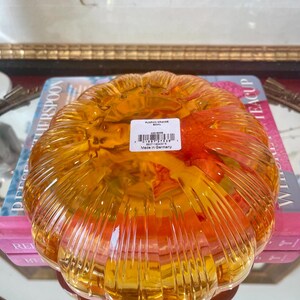 May include: A translucent orange glass bowl shaped like a pumpkin with vertical ribbed detailing. A white rectangular product label is affixed to the center, featuring the text PUMPKIN ORANGE BOWL, a barcode, and the text Made in Germany.