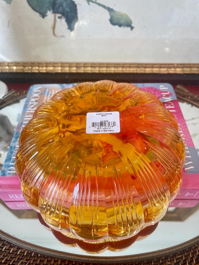 May include: A translucent orange glass bowl shaped like a pumpkin with vertical ribbed detailing. A white rectangular product label is affixed to the center, featuring the text PUMPKIN ORANGE BOWL, a barcode, and the text Made in Germany.
