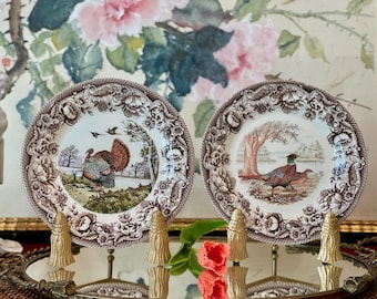 May include: Two round ceramic plates featuring brown transferware floral borders. The left plate displays a turkey in a landscape, while the right plate depicts a pheasant near a tree. Both plates are displayed on gold-toned stands.