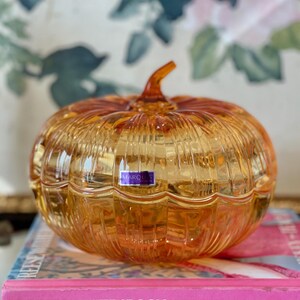 May include: A translucent amber-colored glass pumpkin-shaped lidded dish with vertical ribbed detailing and a small stem on top. A rectangular purple and white sticker with the text MARQUIS is affixed to the front of the glass pumpkin.
