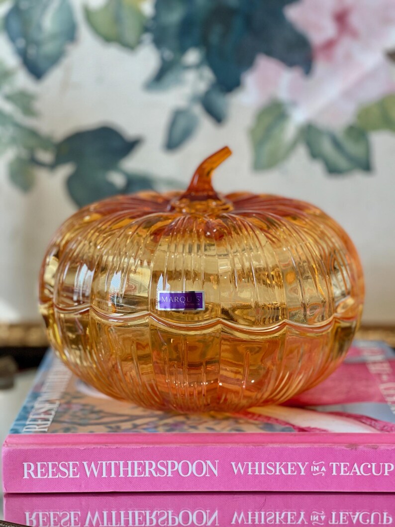 May include: A translucent amber-colored glass pumpkin-shaped lidded dish with vertical ribbed detailing and a small stem on top. A rectangular purple and white sticker with the text MARQUIS is affixed to the front of the glass pumpkin.
