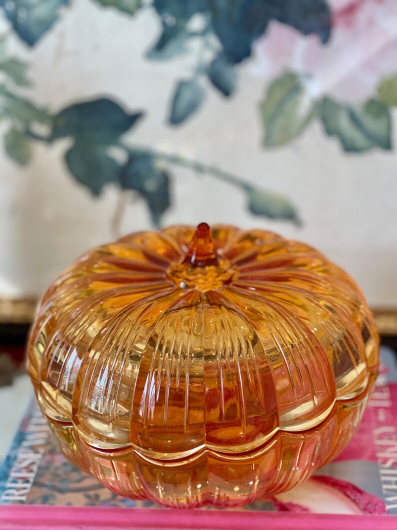 May include: A translucent amber-colored glass pumpkin-shaped container with a lid. The glass features vertical ribbed detailing radiating from a central pointed stem at the top. The object has a rounded, segmented form resembling a gourd.