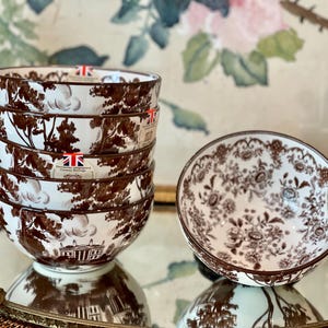 May include: A stack of four ceramic bowls and one separate bowl featuring a brown and white transferware pattern. The exterior displays a landscape with trees and a building, while the interior of the separate bowl shows a floral motif.