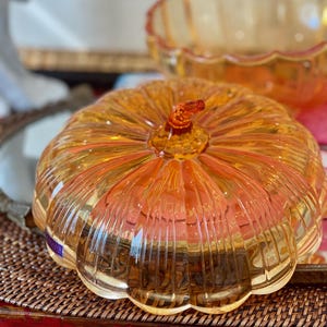 May include: A translucent amber-colored glass pumpkin-shaped lidded dish with vertical ribbed detailing and a small stem handle on top. The lid is rounded and features a scalloped edge, reflecting light through its clear, warm-toned glass surface.