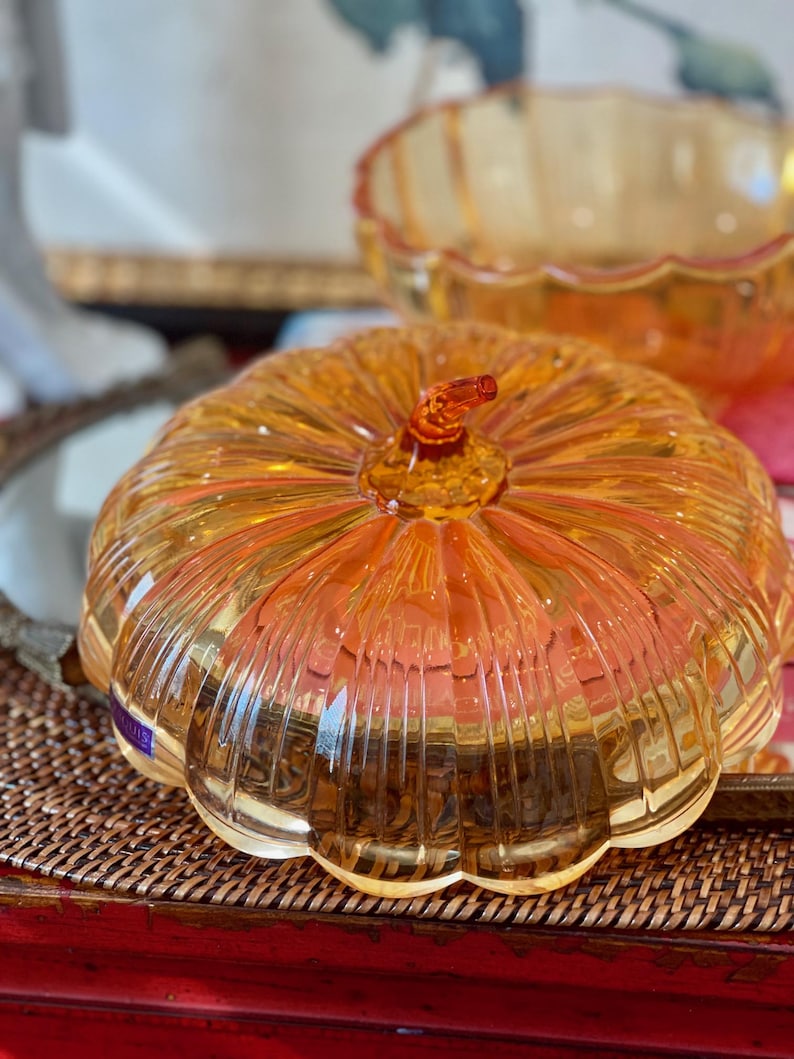 May include: A translucent amber-colored glass pumpkin-shaped lidded dish with vertical ribbed detailing and a small stem handle on top. The lid is rounded and features a scalloped edge, reflecting light through its clear, warm-toned glass surface.