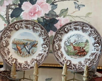 May include: Two round ceramic plates featuring a brown floral border pattern. The left plate displays two ducks in a marshy landscape. The right plate depicts two deer resting in a grassy field with trees. Both plates have a cream-colored center.