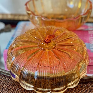 May include: A translucent amber-colored glass pumpkin-shaped lidded dish with vertical ribbed detailing and a small stem handle, accompanied by a matching open glass bowl with a scalloped rim, both featuring a glossy finish and warm orange tones.