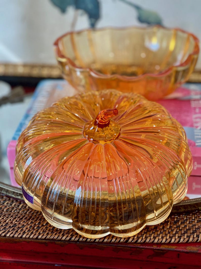 May include: A translucent amber-colored glass pumpkin-shaped lidded dish with vertical ribbed detailing and a small stem handle, accompanied by a matching open glass bowl with a scalloped rim, both featuring a glossy finish and warm orange tones.
